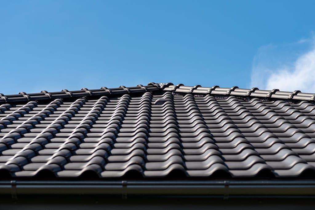 Close-up view of a dark, wavy tiled roof with a clear blue sky in the background. The roof tiles are neatly arranged in rows, and sunlight creates a slight sheen on their surface.