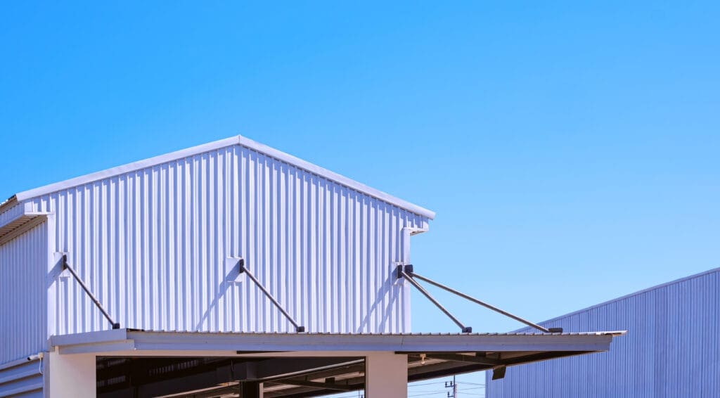 A close-up view of a modern industrial building with a corrugated metal exterior, set against a clear blue sky. Diagonal support beams are visible on the structure’s roof.