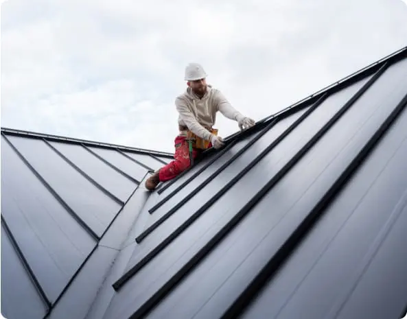 Roofing contractor wearing a white hard hat and safety harness installs standing seam metal panels on a residential roof under cloudy skies