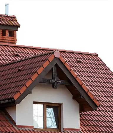 Steeply pitched residential roof with reddish‑brown ceramic tiles, dormer window, and brick chimney under clear skies